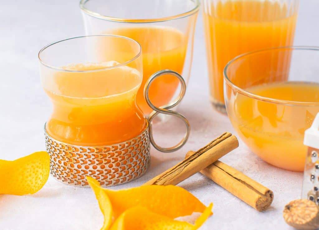 Four various glasses filled with mulled apple juice, some orange peels curled, cinnamon sticks and a piece of whole nutmeg with a small nutmeg grater on a pale grey background.