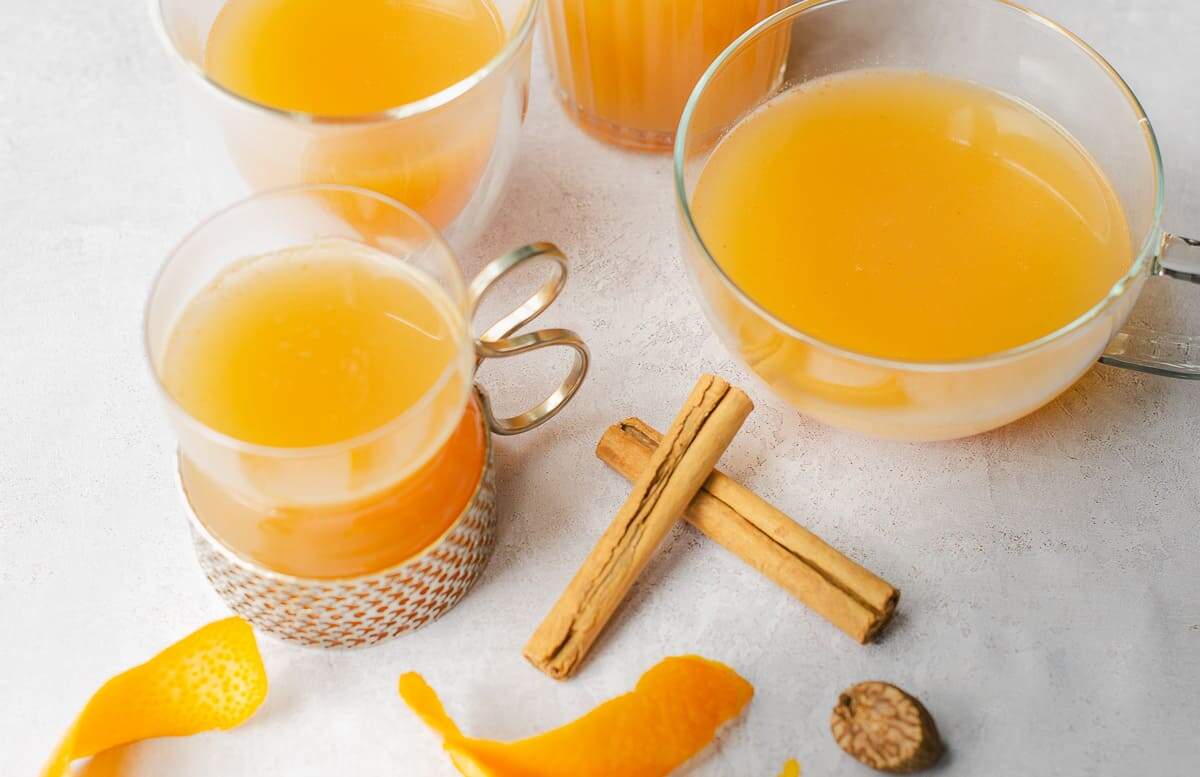 Four different shaped glass mugs and glasses filled with warm mulled apple juice, some twisted orange peels, some cinnamon sticks and a small grater with a piece of whole nutmeg.