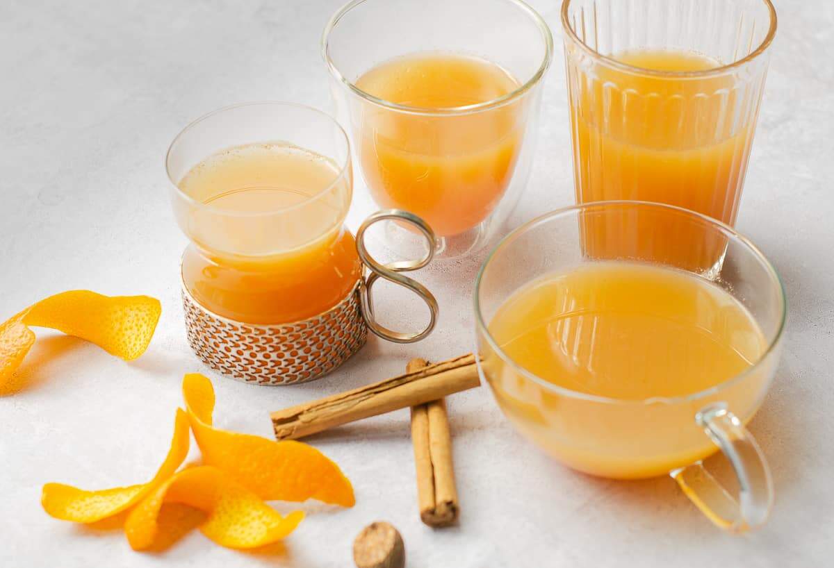 Various glasses filled with mulled apple juice, some orange peels curled, cinnamon sticks and a piece of whole nutmeg with a small nutmeg grater on a pale grey background.