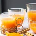 Various glasses filled with mulled apple juice, some orange peels curled, cinnamon sticks and a piece of whole nutmeg on a pale grey background.