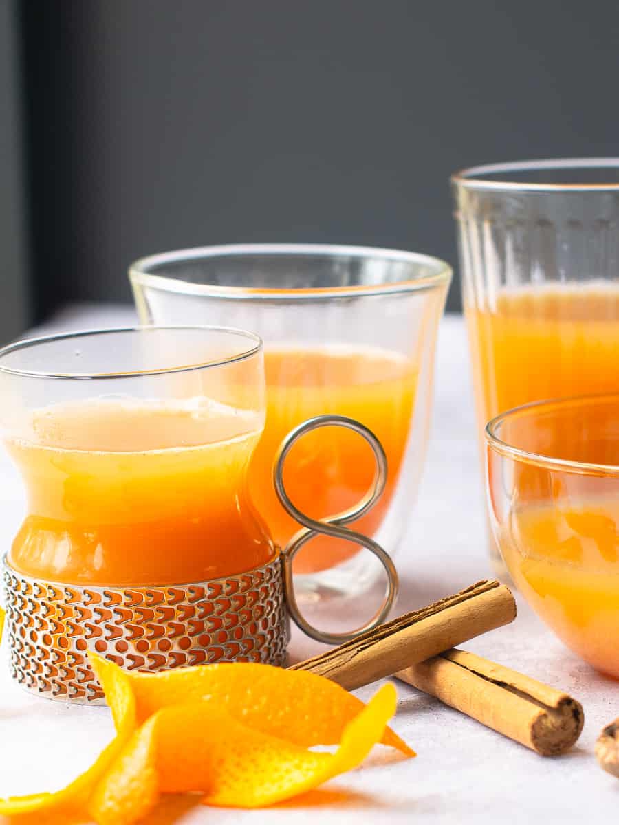 Various glasses filled with mulled apple juice, some orange peels curled, cinnamon sticks and a piece of whole nutmeg on a pale grey background.