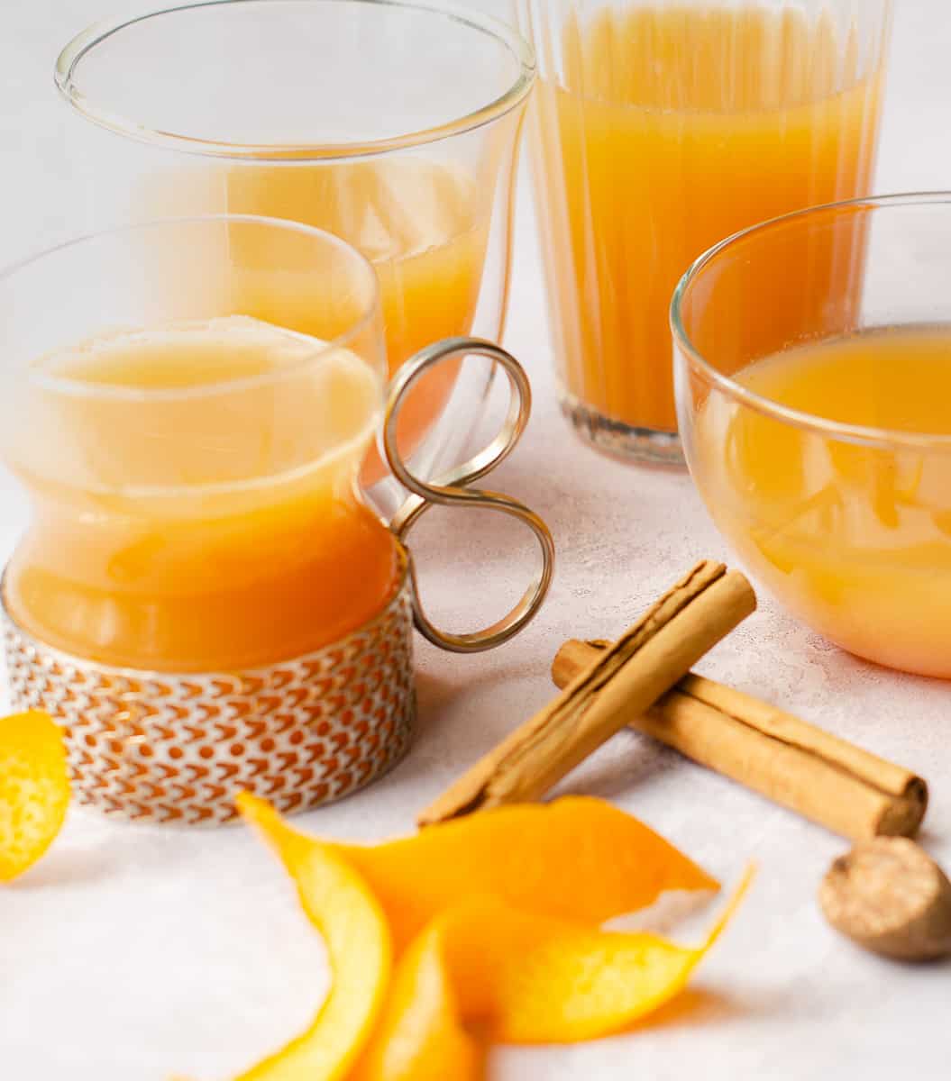 Various glasses filled with mulled apple juice, some orange peels curled, cinnamon sticks and a piece of whole nutmeg on a pale grey background.