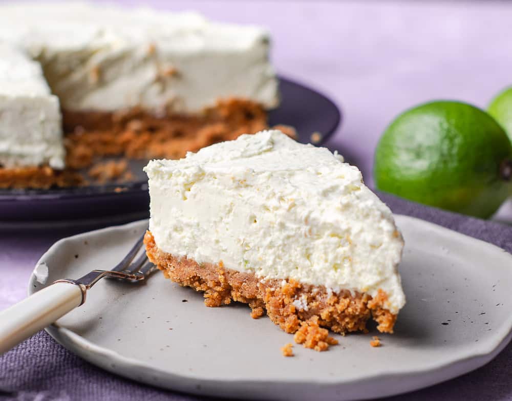 A closeup of a slice of no bake lime cheesecake from a top view position on a grey ceramic plate sitting on a dark blue napkin and the rest of the cheesecake to the back with some limes to decorate.