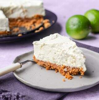 A slice of bake lime cheesecake on a grey ceramic plate sitting on a dark blue napkin and the rest of the cheesecake to the back with some limes to decorate.