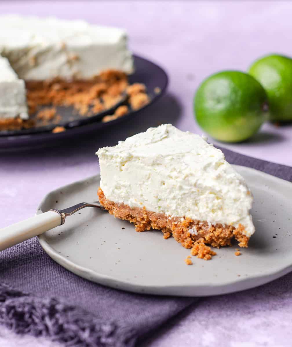 A slice of bake lime cheesecake on a grey ceramic plate sitting on a dark blue napkin and the rest of the cheesecake to the back with some limes to decorate.