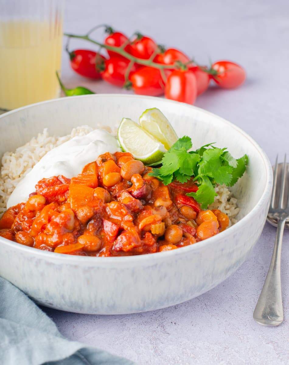 A bowl of vegan bean chilli with basmati rice and topped with sour cream. Some vine tomatoes to the back and a drink on a grey backdrop with a pale blue napkin and cutlery to the sides of the bowl