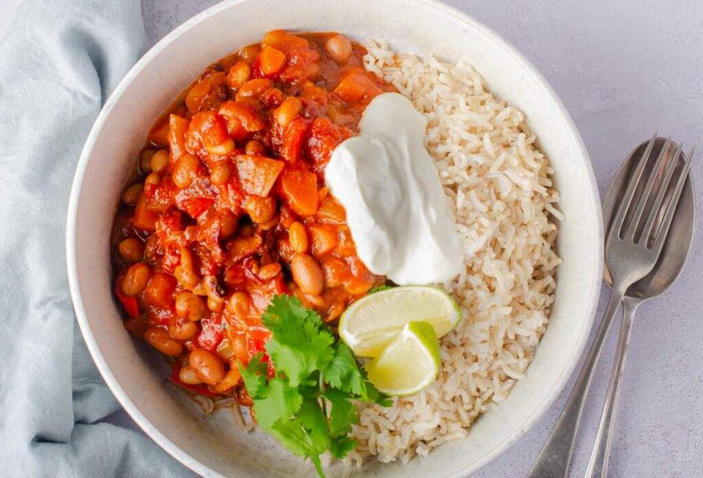 A top down view of a bowl of vegan bean chilli with rice and soured cream garnished with coriander and limes in a pale blue mottled bowl on a grey backdrop and a fork and spoon to the right.