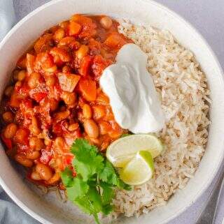 A top down view of a bowl of vegan bean chilli with rice and soured cream garnished with coriander and limes in a pale blue mottled bowl on a grey backdrop and a fork and spoon to the right.