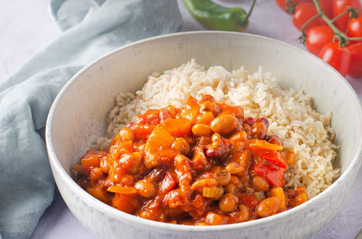 A pale blue mottled ceramic bowl filled with vegan bean chilli with basmati rice some vine tomatoes to the side back and a pale blue napkin to the left sitting on a grey surface.