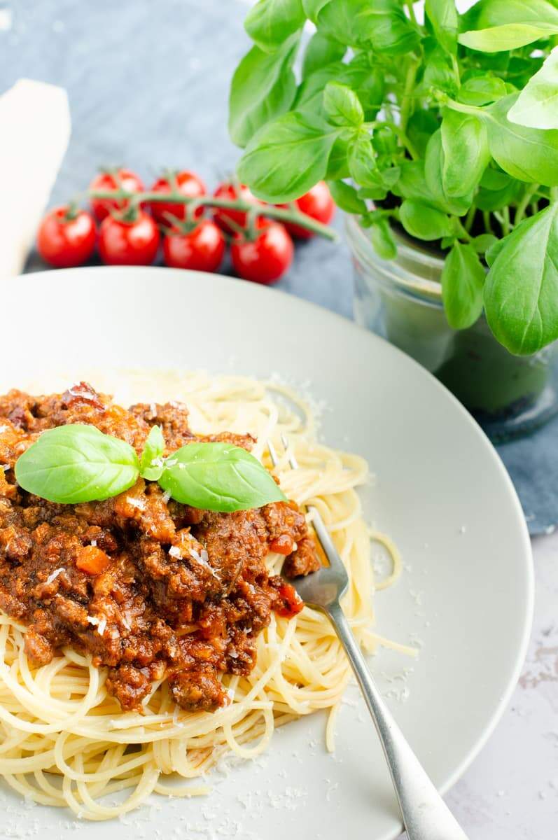 An enticing plate of Spaghetti Bolognese served ready on a grey plate with a fork and a vibrant basil plant in the background along with a bunch of vine tomatoes
