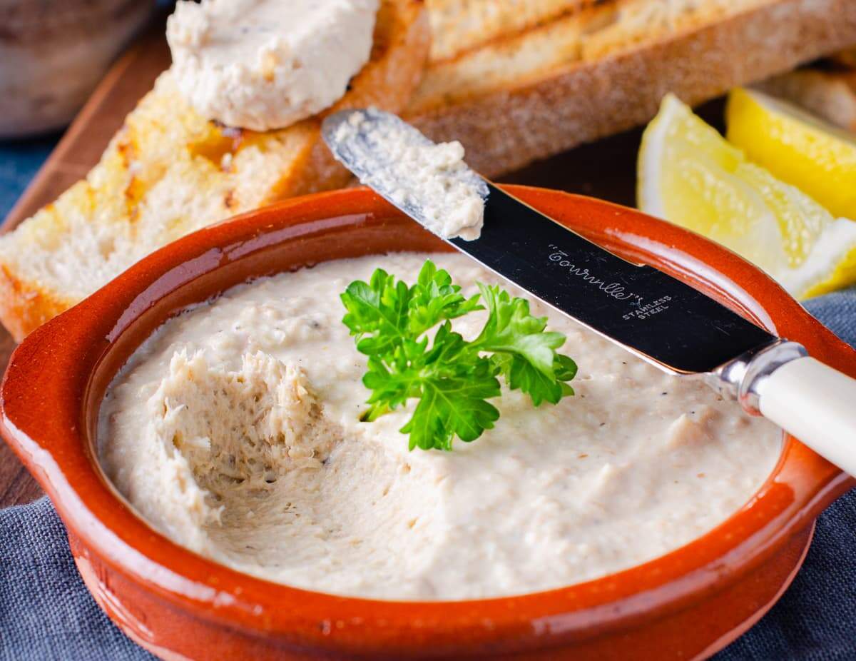 a closeup view of a dish of smoked mackerel pate garnished with parsley and sitting o a blue napkin with a bone handle knife taking a first dip.