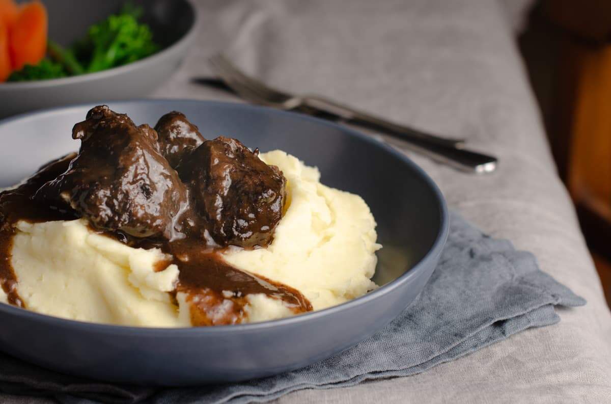 A table setting of a grey linen table cloth, dark blue napkins with a blue bowl filled with creamy mashed potatoes and topped with slow braised beef cheeks topped with a rich thick gravy and some vegetables to the back.