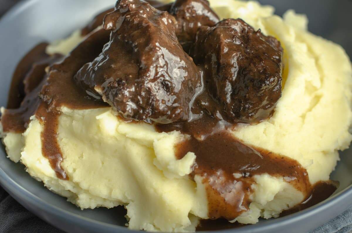 A closeup preview of a blue bowl containing creamy buttery mashed potatoes and topped with slow braised beef cheeks and rich gravy.