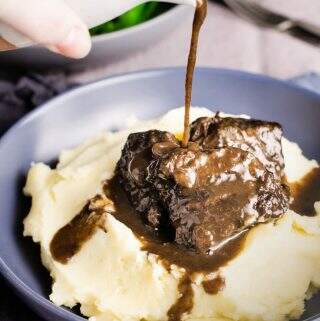 A blue bowl of buttery mashed potato topped with slow braised beef cheeks and gravy being poured over the top.