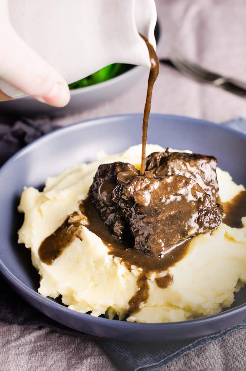 A blue bowl of buttery mashed potato topped with slow braised beef cheeks and gravy being poured over the top.