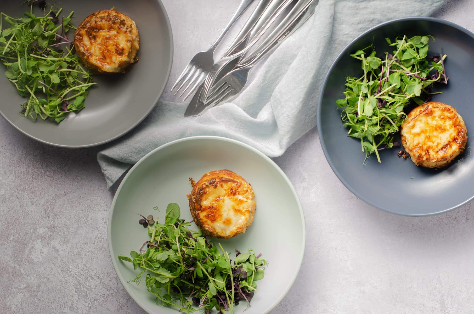Looking above a table set for 3 plates of twice baked cheese souffles and green salads on plates of green, blue and grey and a napkin with cutlery.