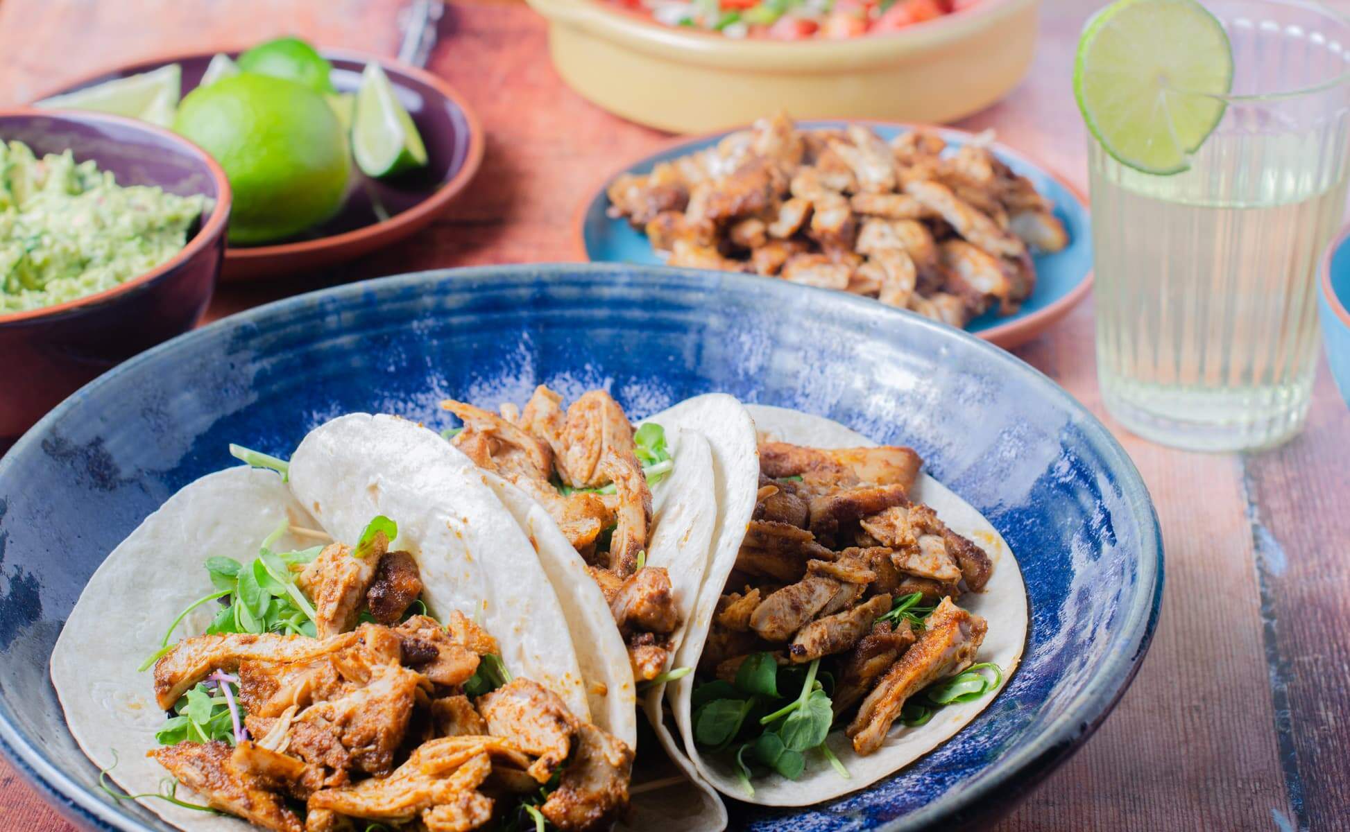 A table setting for a taco party with chicken tacos on a blue platter, lime cordial drinks in the back along with salsa and guacamole and more limes.