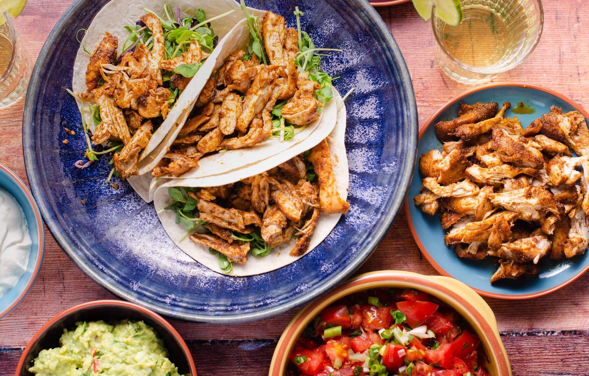 a vibrant table setting for a taco party including chicken tacos on a bright blue platter, salsa, guacamole and more shredded chicken.