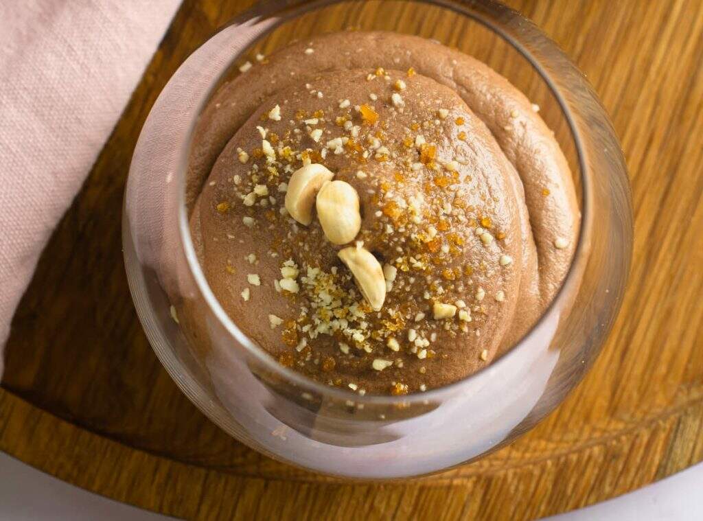 Looking into a glass of nutella mousse topped with praline and whole hazelnuts served on a wooden tray and with a pink napkin.