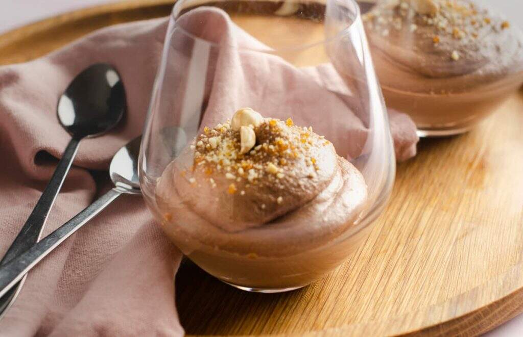 Close of of 2 glasses of Nutella Mousse on a wooden serving tray decorated with praline and whole hazelnuts with spoons and a pink linen napkin