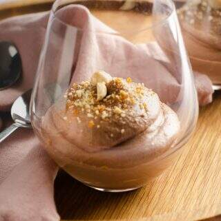 Close of of 2 glasses of Nutella Mousse on a wooden serving tray decorated with praline and whole hazelnuts with spoons and a pink linen napkin