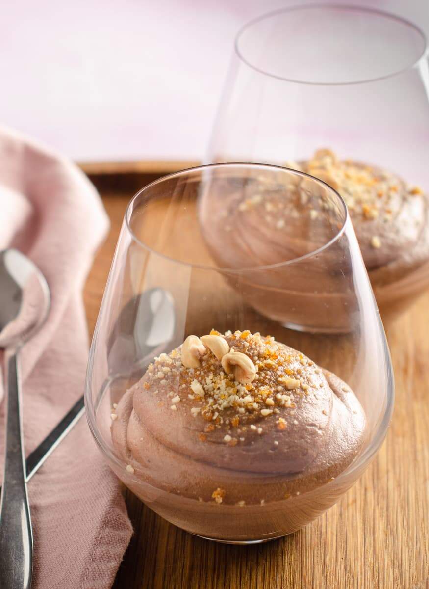 Glasses served with Nutella mousse inside garnished with a praline crumb & whole hazelnuts on a wooden board and a pink napkin and spoons to the left.