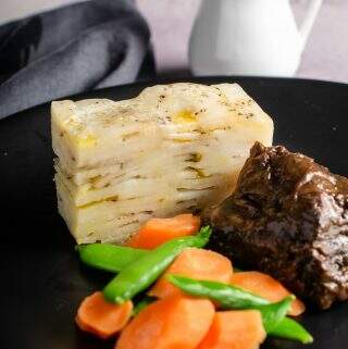 Pave potatoes sliced and served with beef cheek stew and fresh vegetables on a black plate and a pot of gravy to the back.