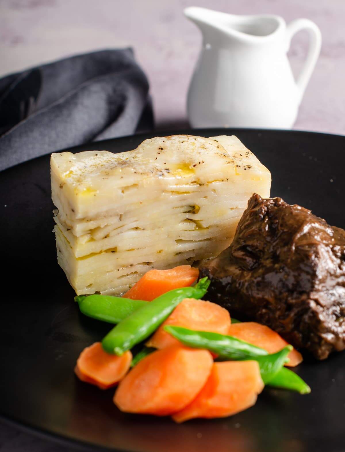 Pave potatoes sliced and served with beef cheek stew and fresh vegetables on a black plate and a pot of gravy to the back.