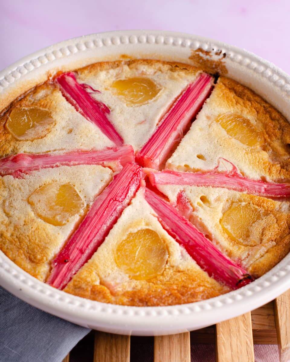 Rhubarb clafoutis with stem ginger, decorated in a cream serving dish with pink rhubarb stems and slices of stem ginger in a star shaped pattern.