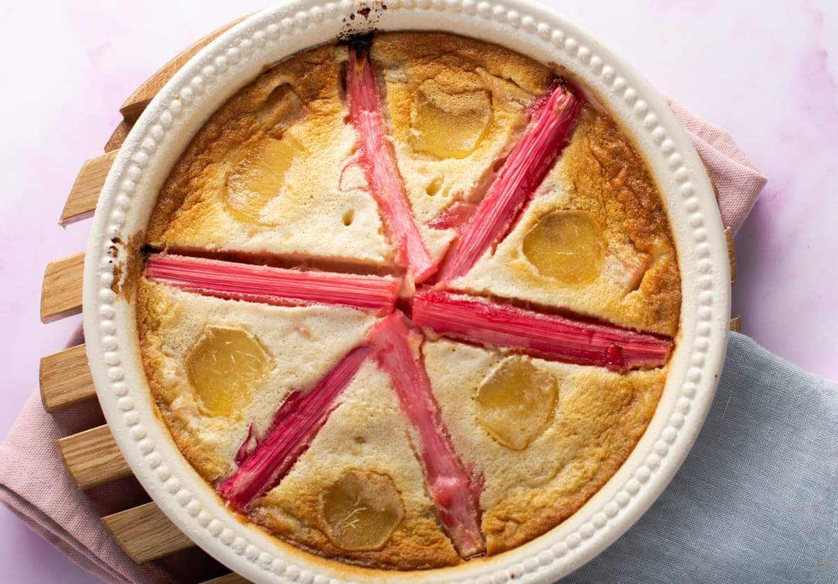 Rhubarb clafoutis with stem ginger, a deliciously light dessert sitting on a wooden trivet and decorated with a star shaped pattern with stems of poached pink rhubarb.