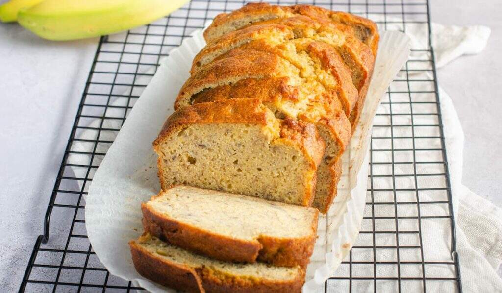 A freshly baked banana bread sliced, sitting on the parchment paper on a black cooling rack and some fresh bananas in the back.
