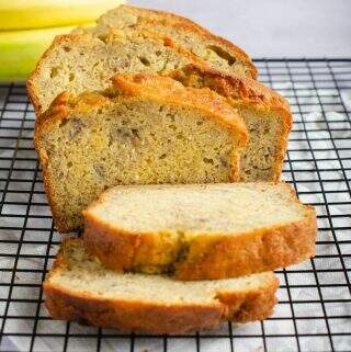 Front view of a sliced banana loaf (bread) on a wire cooling rack and some fresh bananas to the back.