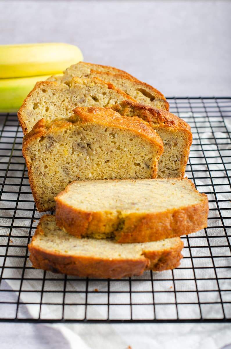 Front view of a sliced banana loaf (bread) on a wire cooling rack and some fresh bananas to the back.
