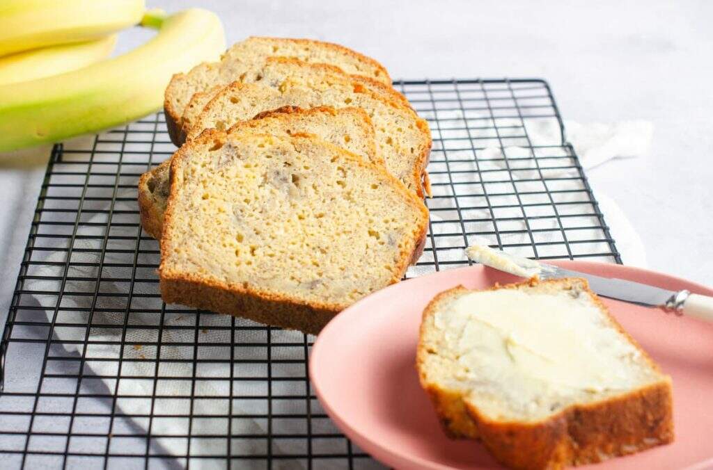 A buttered slice of banana bread in the foreground on a pink plate and the rest of the loaf to the back sitting on a wire cooling rack with some fresh bananas to the back.