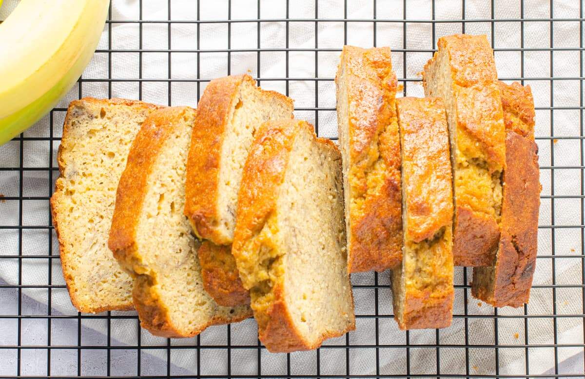 8 perfectly sliced pieces banana bread from above laid out on a black wire cooling rack with some fresh bananas to the side, ready to be eaten.