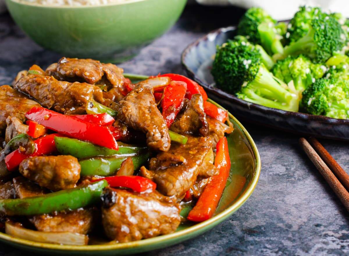 A table set with a dish of beef and black bean sauce to the front, stir fried broccoli to the back and a pair of chopsticks on a dark marbled table.