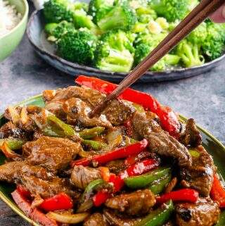 A dish of beef and black bean sauce with a hand using chopsticks to lift some of the dish with broccoli to the back and a bowl of rice.