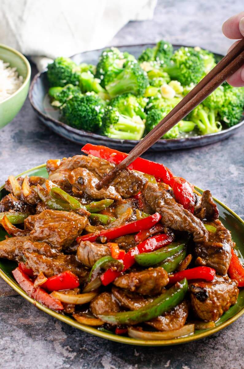 A dish of beef and black bean sauce with a hand using chopsticks to lift some of the dish with broccoli to the back and a bowl of rice.