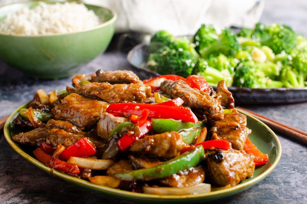 A dish of beef and black bean sauce with chopsticks to the side on a green plate and a dark marbled surface and a dish of stir fried broccoli to the back along with a bowl of rice.