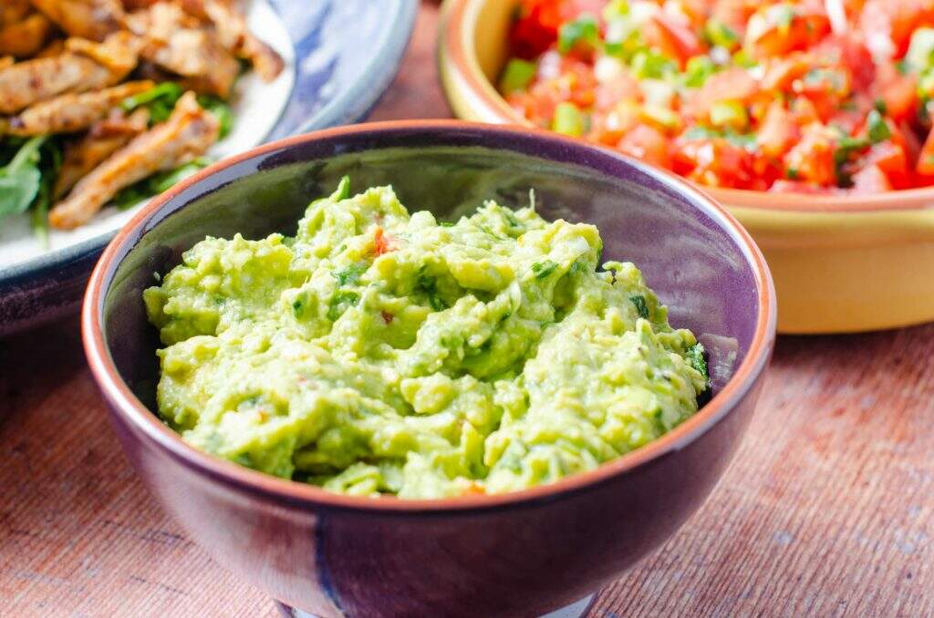 A bowl of freshly made guacamole to the front with fresh salsa and chicken tacos to the back all on a dark wooden surface.