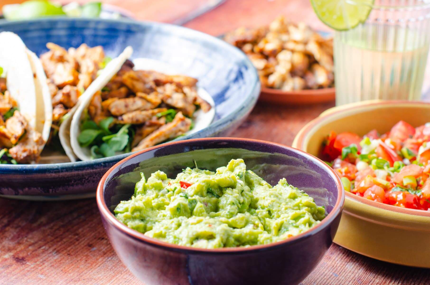 A dark wooden table set for a taco party with fresh guacamole in a purple bowl, fresh salsa to the side, chicken tacos made up and lime drinks in the back.