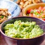 Super fresh guacamole in. purple bowl with all the elements for a taco party in the back, chicken, salsa, wraps all on a wooden table.