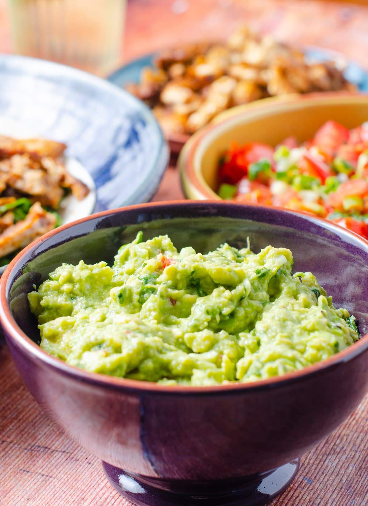 Super fresh guacamole in. purple bowl with all the elements for a taco party in the back, chicken, salsa, wraps all on a wooden table.