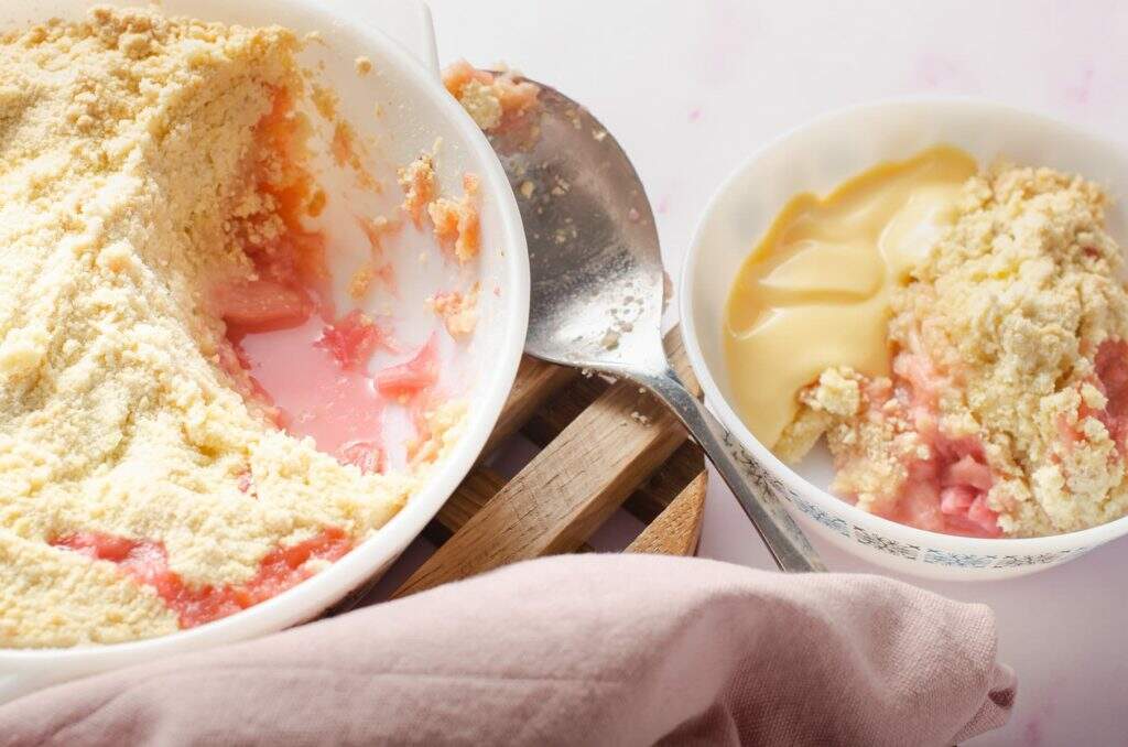 A family serving dish of rhubarb crumble on the left, a serving spoon and a bowl of rhubarb crumble with a portion of custard all served with a pik napkin to the front.