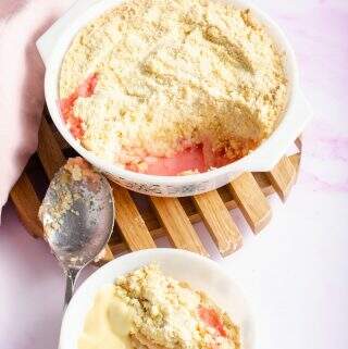 Rhubarb crumble with a portion taken out and sitting in a bowl with custard, a pink napkin to the back and the serving dish on a wooden trivet.