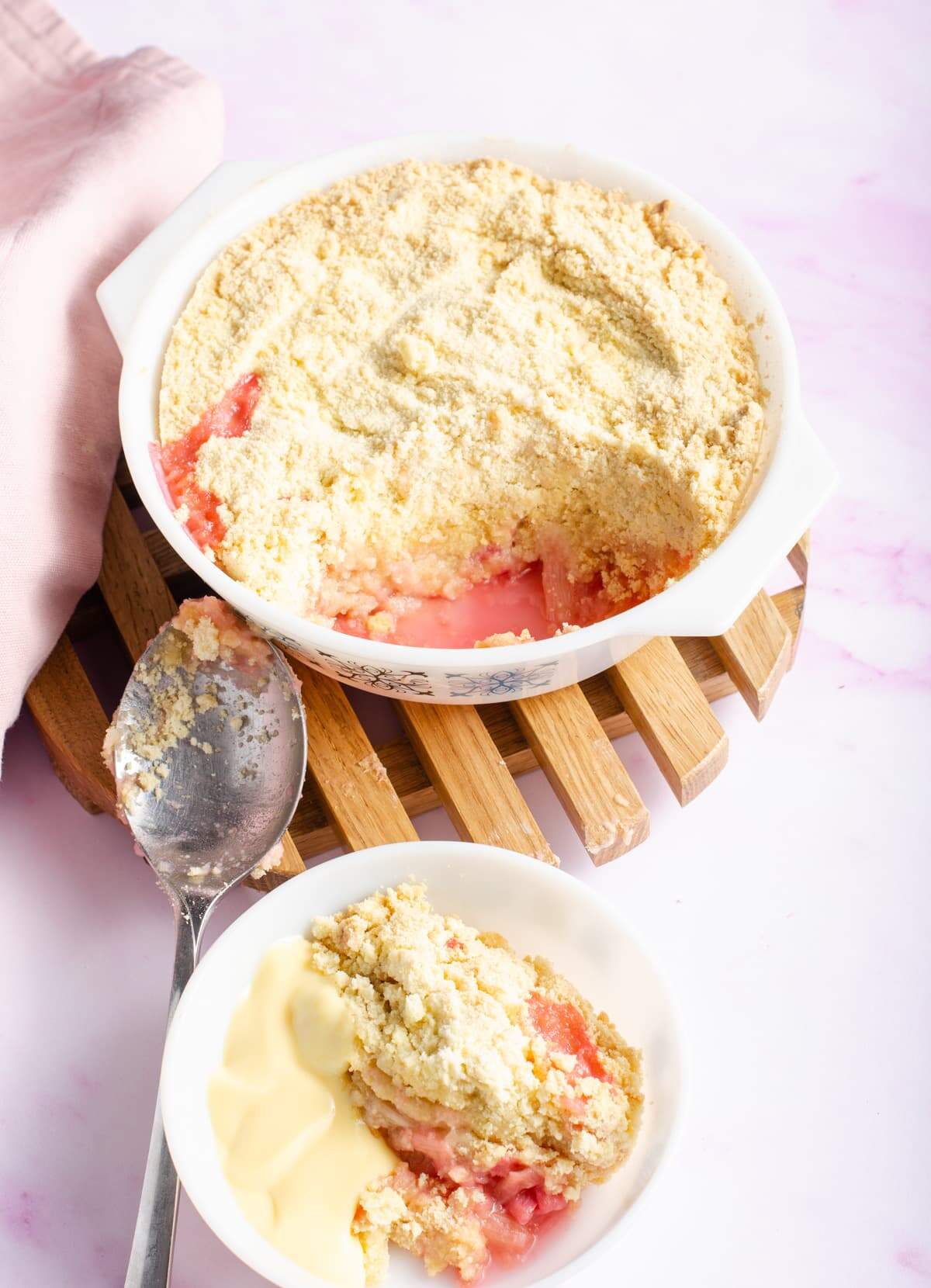 Rhubarb crumble with a portion taken out and sitting in a bowl with custard, a pink napkin to the back and the serving dish on a wooden trivet.