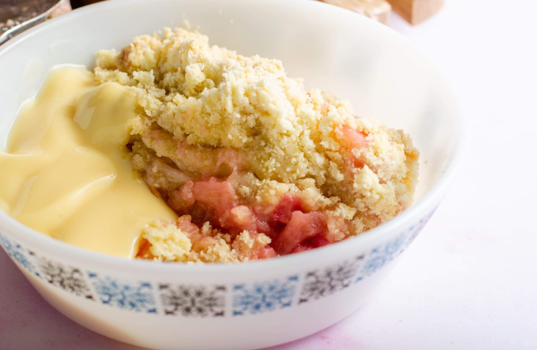 A served portion of rhubarb crumble in a blue & white bowl and a generous portion of custard served with it.