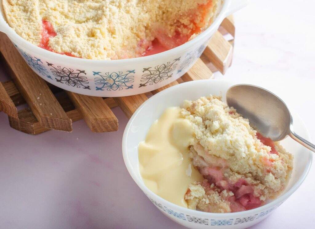 Rhubarb crumble with a portion taken out and sitting in a bowl with custard, a pink napkin to the back and the serving dish on a wooden trivet.