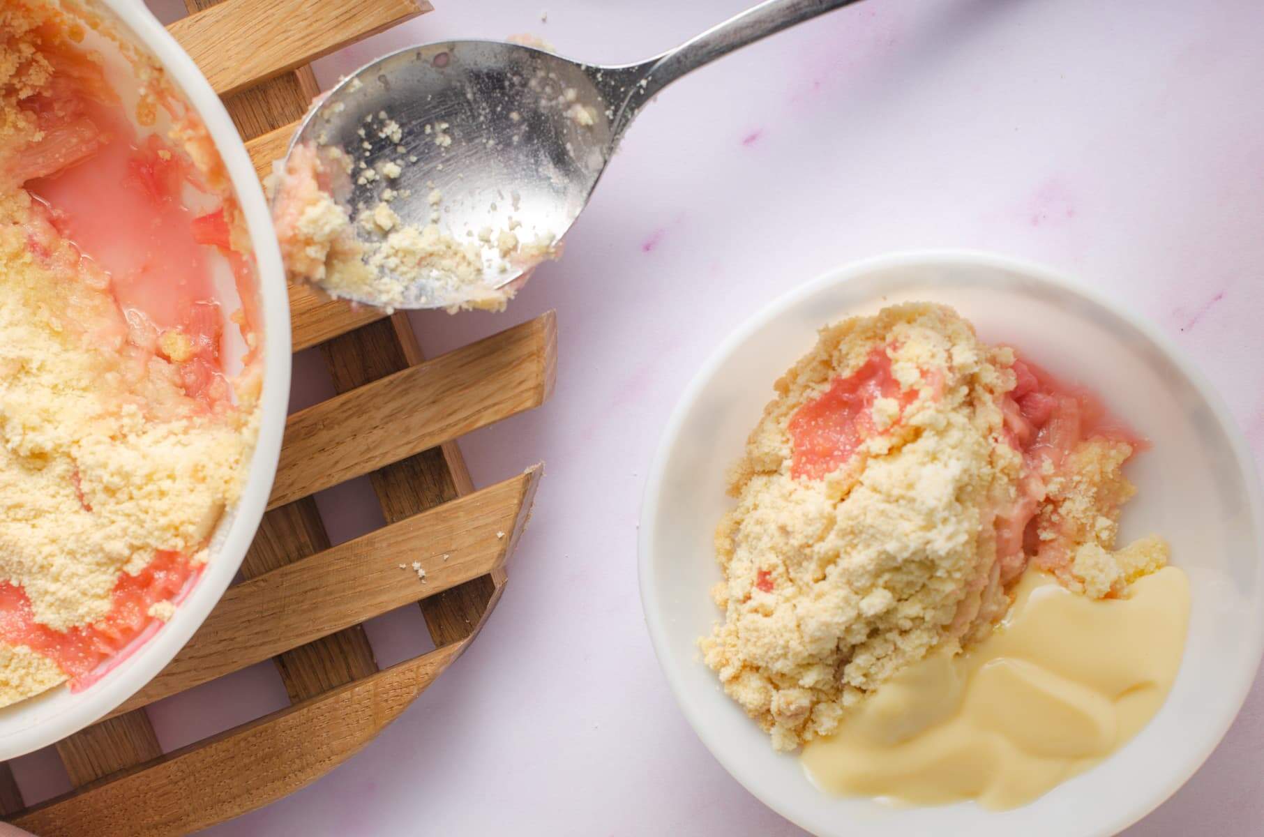 Rhubarb crumble with a portion taken out and sitting in a bowl with custard, a pink napkin to the back and the serving dish on a wooden trivet.