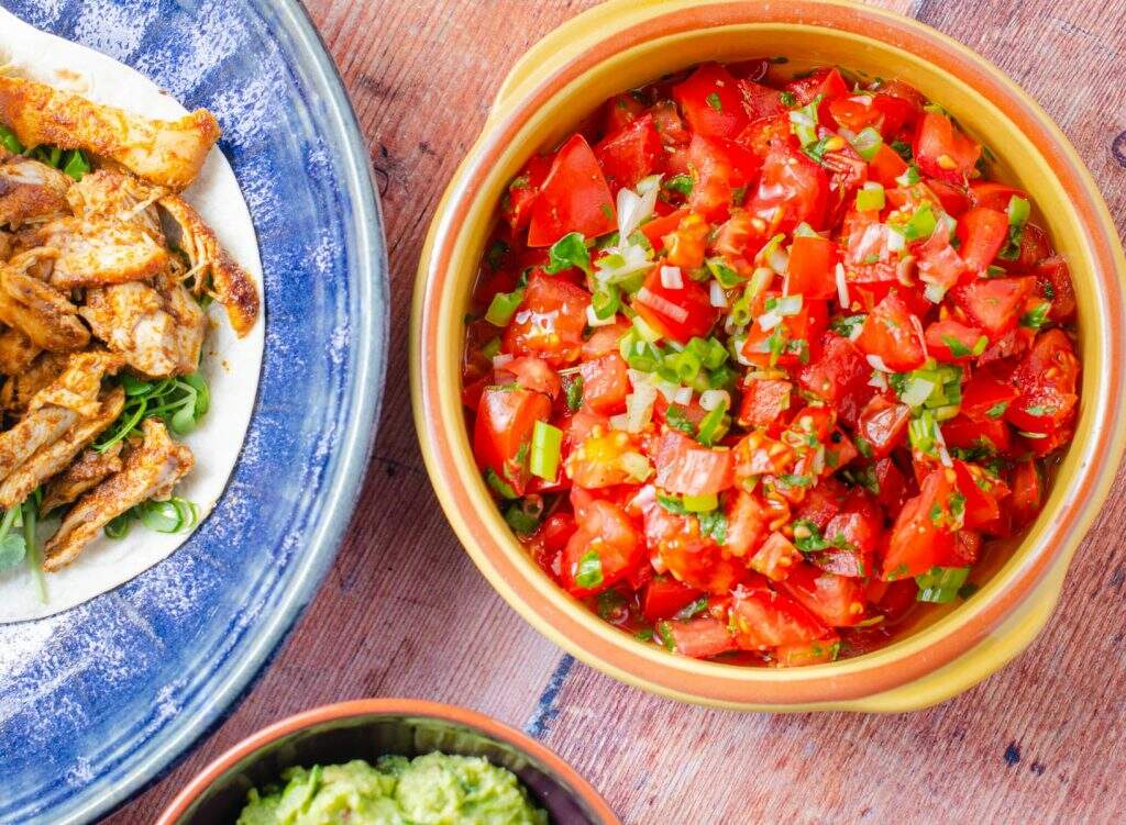 Fresh tomato salsa in a yellow bowl from above with chicken tacos on a large blue platter to the side and a bowl of fresh guacamole to the bottom all against a dark wooden backdrop.